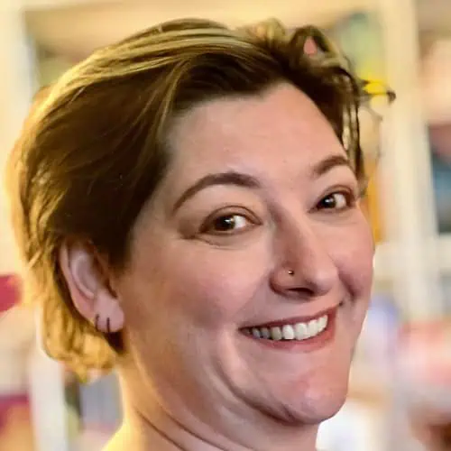 Headshot of Emma Leadley smiling at the camera. They have multiple piercings, no makeup and short brown hair.